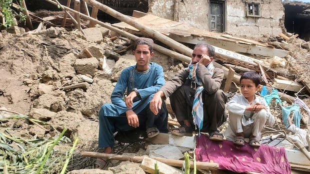 A somber scene of three people sitting amidst rubble from a collapsed building. They appear contemplative and affected by the devastation surrounding them.