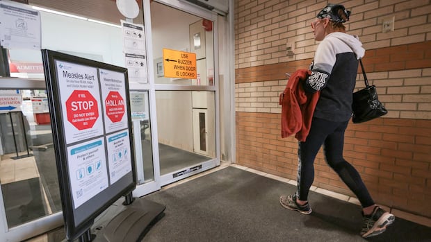 A person walks past a measles alert public health sign, toward a sliding glass door.