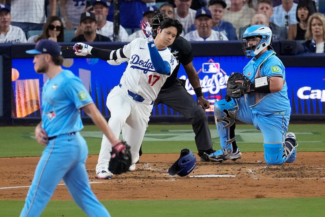 A baseball player swings, misses, and loses his helmet in front of the catcher and umpire. The pitcher waits on the mound.