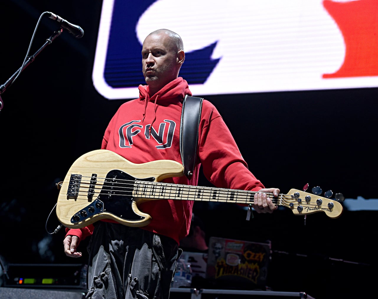 A bald man in a red sweatshirt stands on stage at night with a bass guitar on his shoulder. 