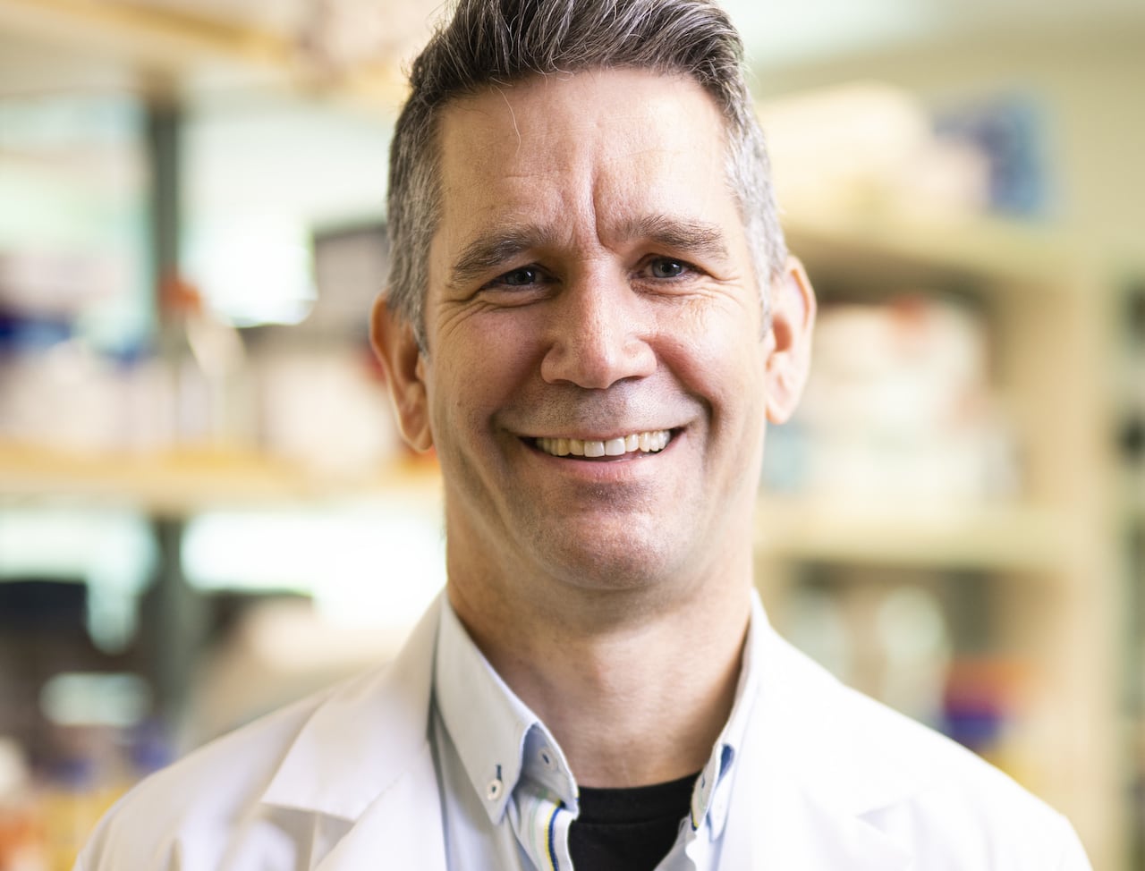 Un homme souriant, portant une chemise à col et une blouse de laboratoire.