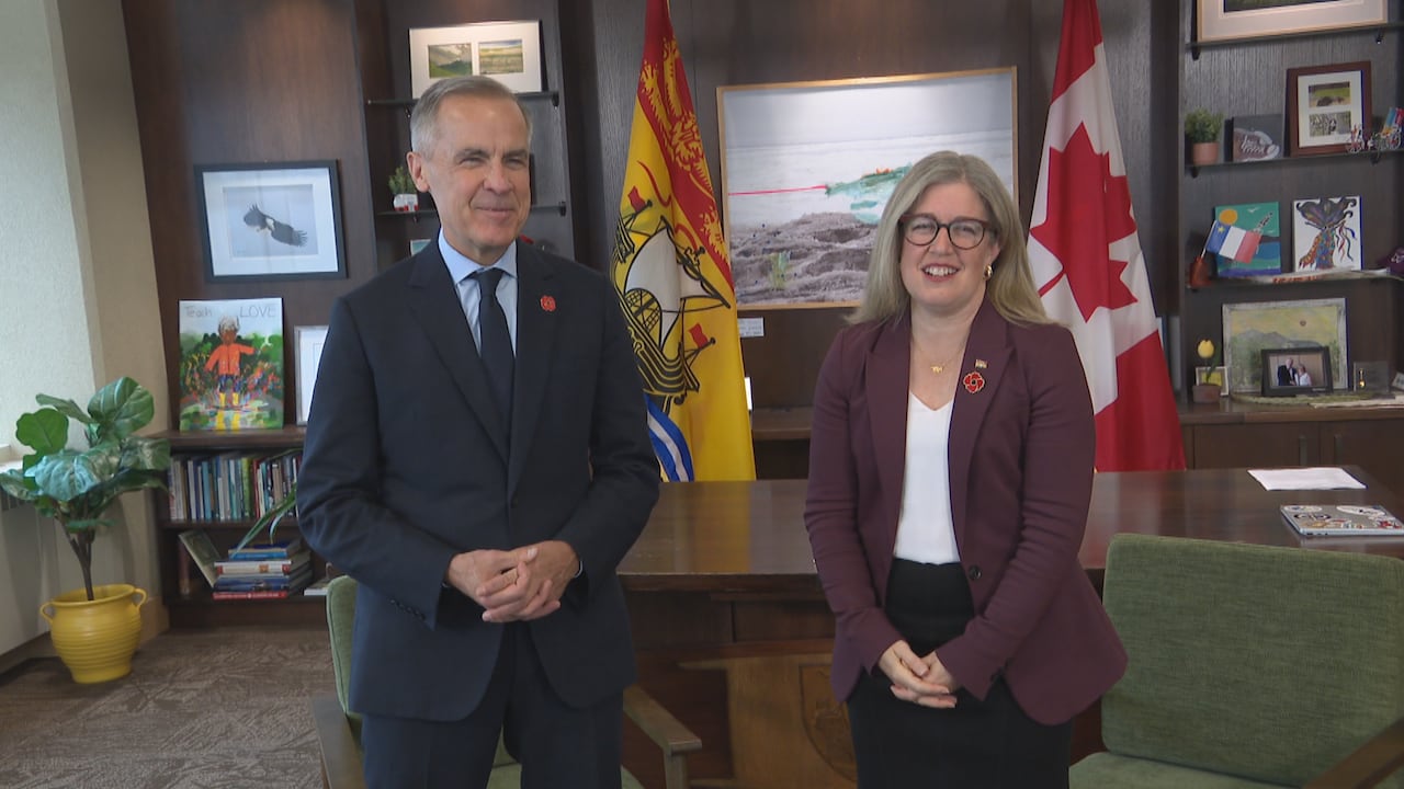 A man in a suit, Mark Carney, and Susan Holt, a woman in a blazer stand side by side in front of a desk.