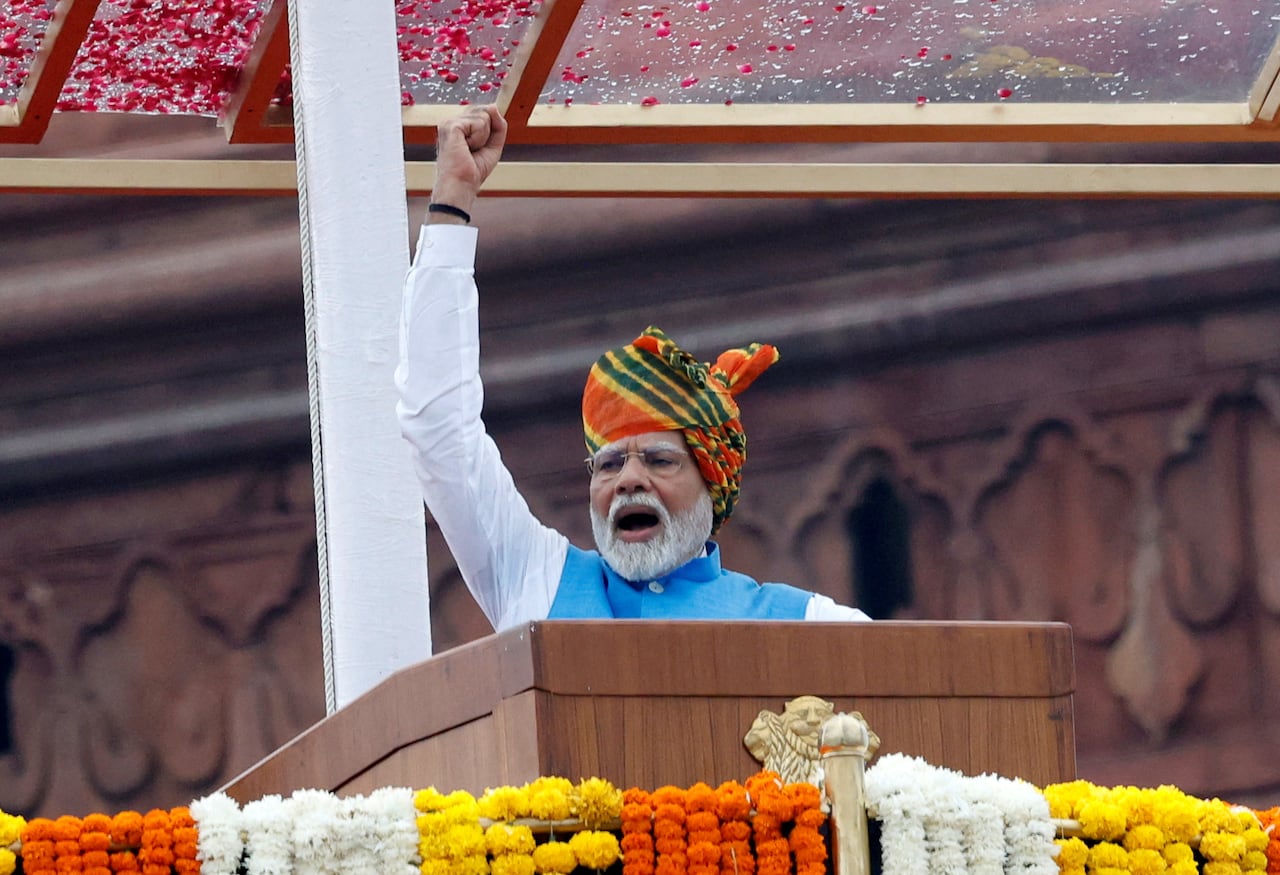 A man in a blue-and-white shirt and multicoloured headdress raises his arm while speaking at a podium outside.