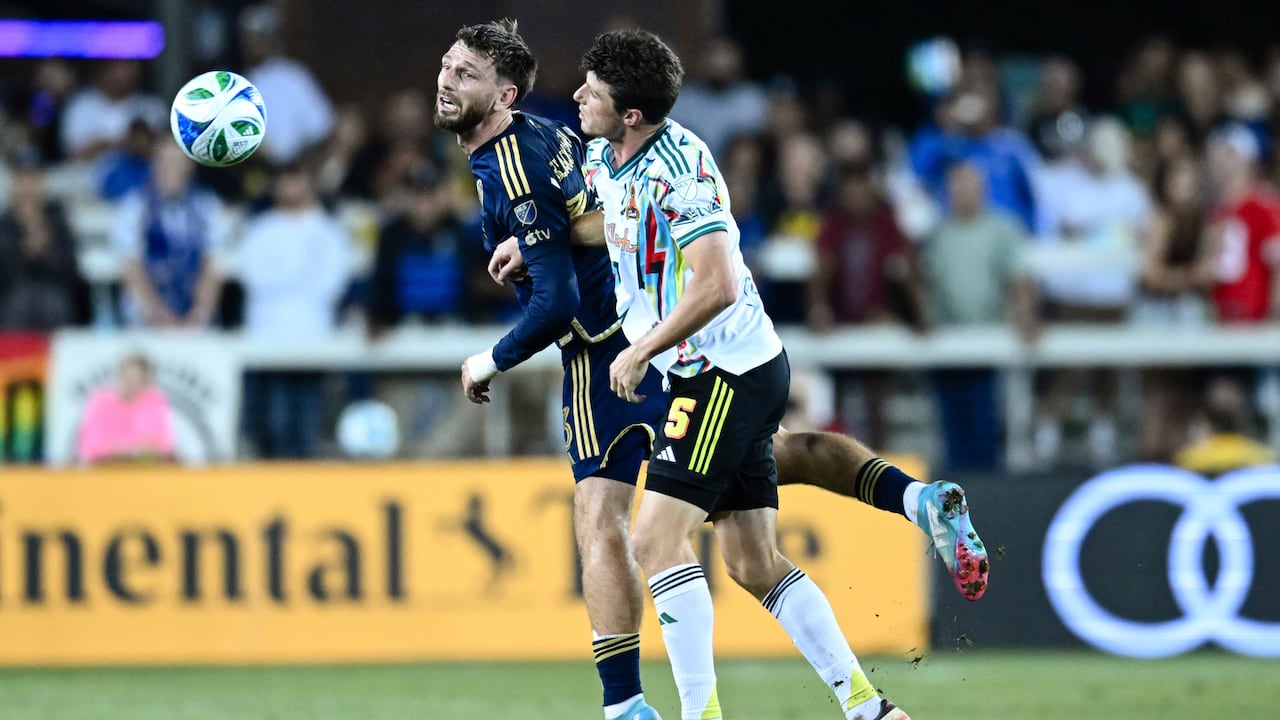 Two soccer players, one in a blue uniform, another in a white uniform, come together as a ball floats through the air.