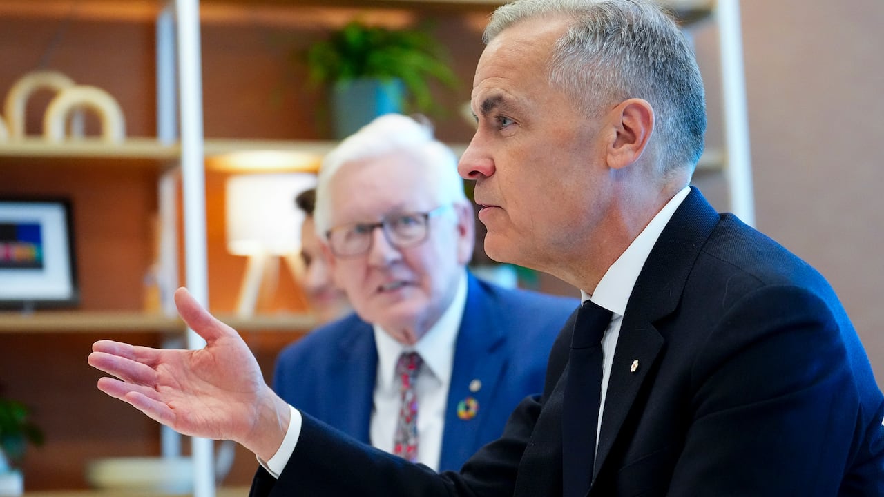 A man gestures while talking in front of another man.