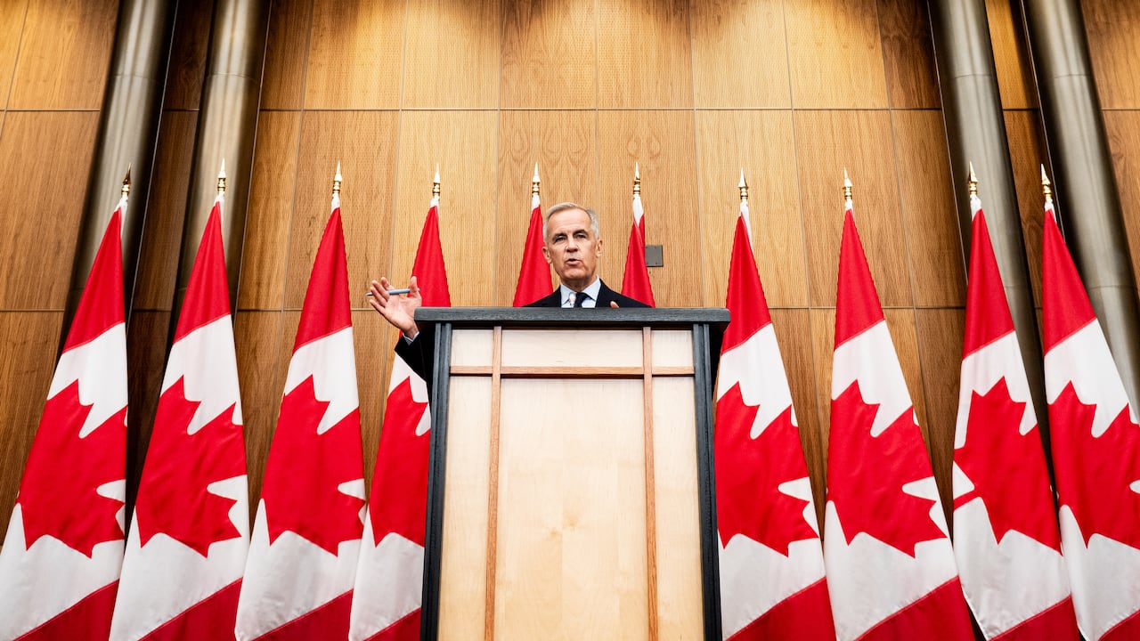 A man standing at a podium, waving one hand, with Canadian flags for a backdrop.