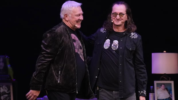 Two men in dark graphic shirts and dark pants stand on stage with mics on their ears, smiling and embracing each other with one arm.