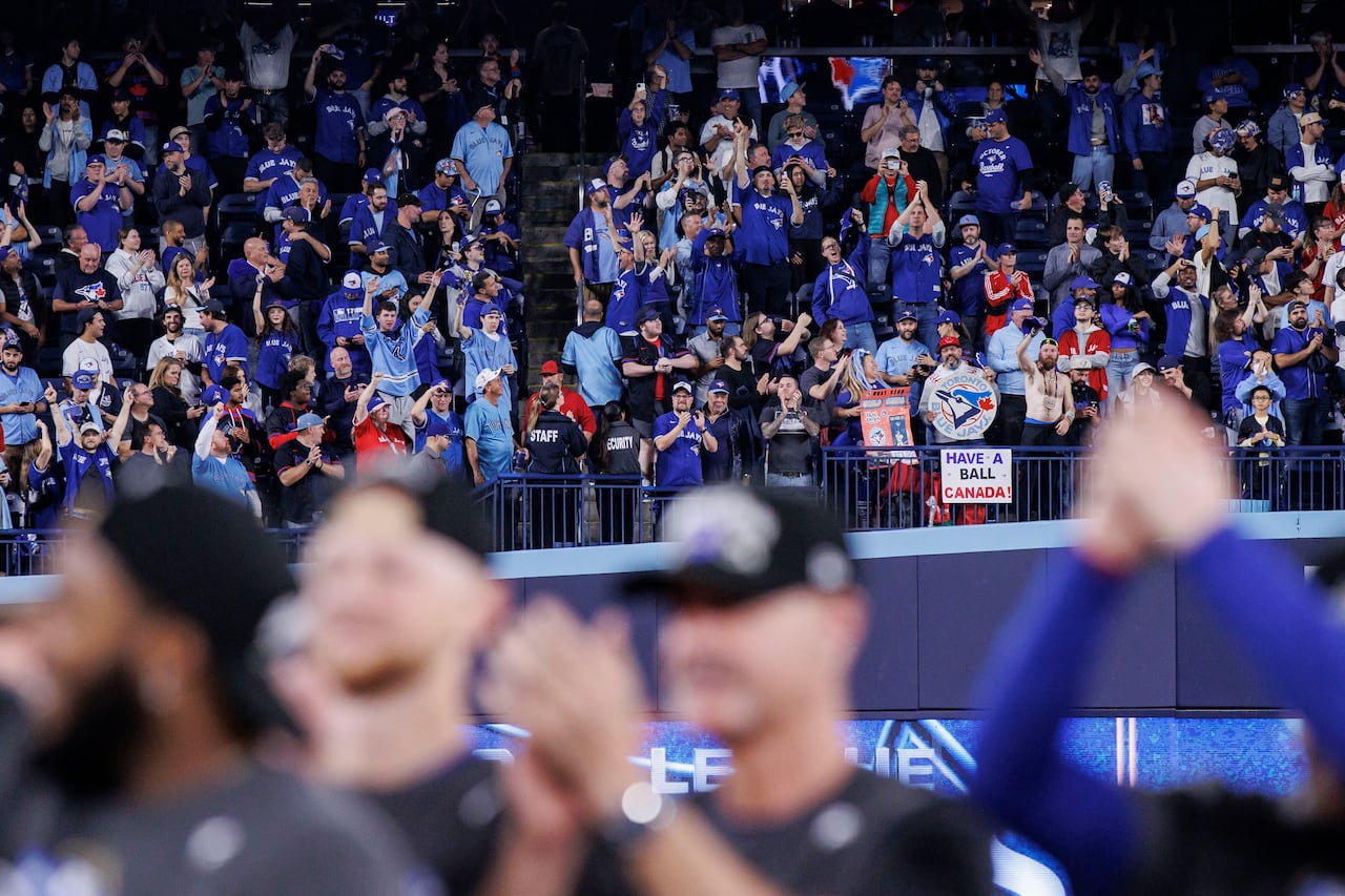 At Rogers Centre, in the foreground, blurry players faces. In the background, fans in the outfield seats cheer on the victory celebration. A sign in the crowd says 'have a ball Canada'