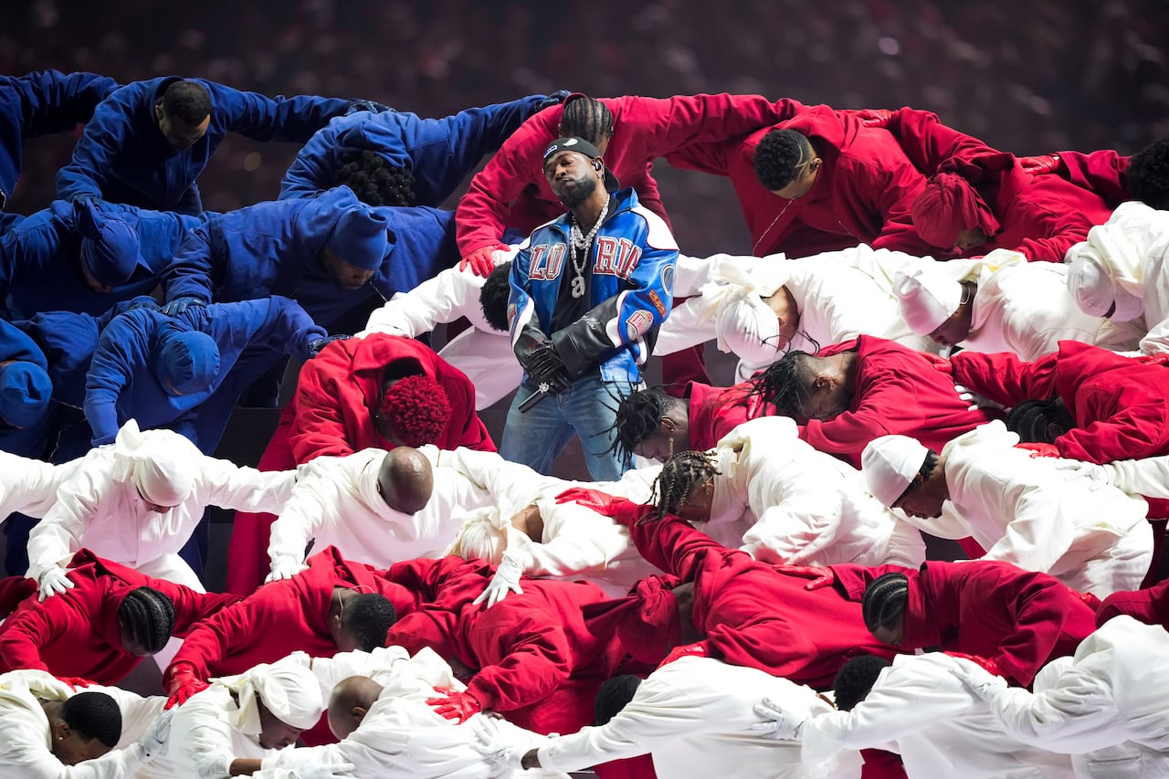 A man wearing a blue leather jacket and jeans stands proudly with his arms crossed in front of him, holding a microphone. Around him are dancers dressed in red, white and blue tracksuits.