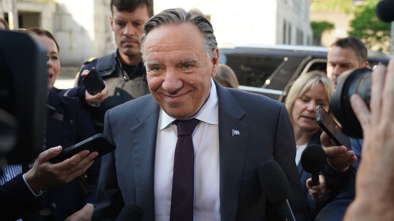 A smiling Francois Legault walking trhough a crowd of reporters on the sidewalk near the national assembly