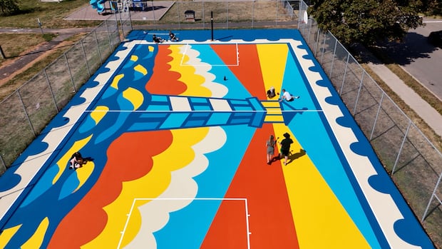 An aerial shot shows five people painting various locations on a basketball court with a design of a large lighthouse shedding beams.