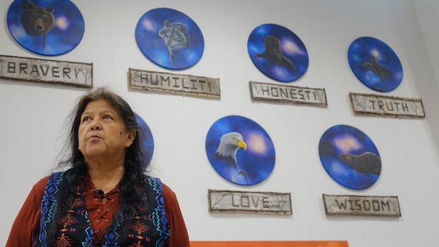  A woman with brown hair wearing a colourful vest is pictured in front of a wall with hangings depicting teachings of love, wisdom, truth, honesty, humility and bravery