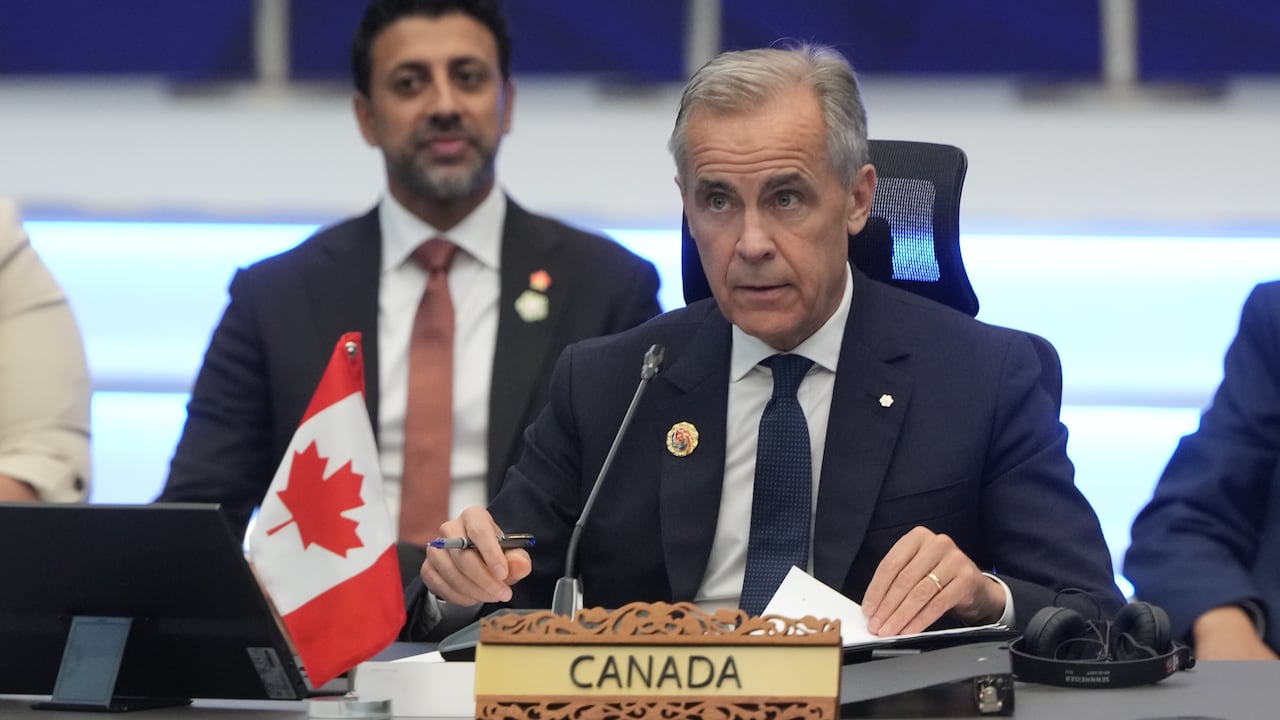 A grey-haired man, wearing a navy suit and tie with a white shirt, sits a table behind a microphone and a wooden nameplate that says "Canada."