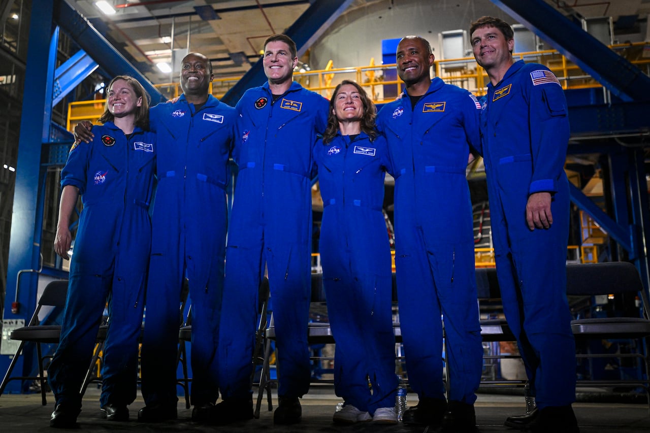 Six people stand hugging each other. They are dressed in matching blue overalls and smile at the camera.