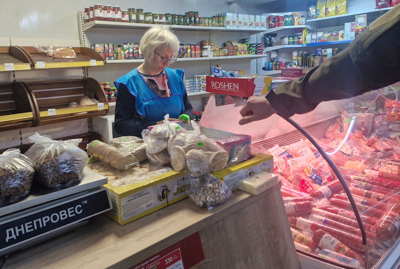 A woman wearing a blue smock stands behind a grocery counter holding bread, beans and meat.