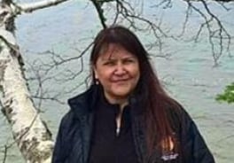 A woman with long black hair stands in front of a lake and a birch tree
