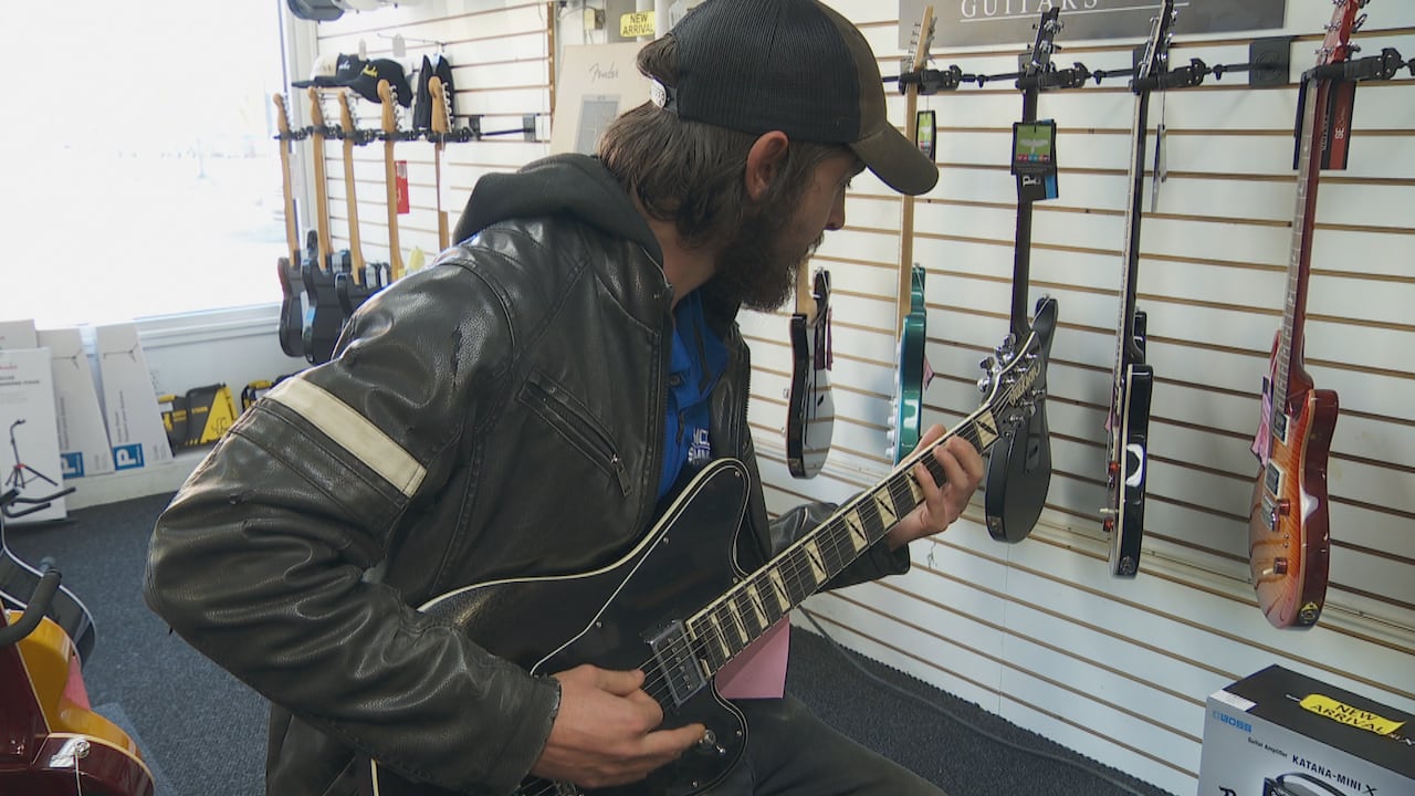 A man wearing a black leather jacket plays a guitar in a music store.