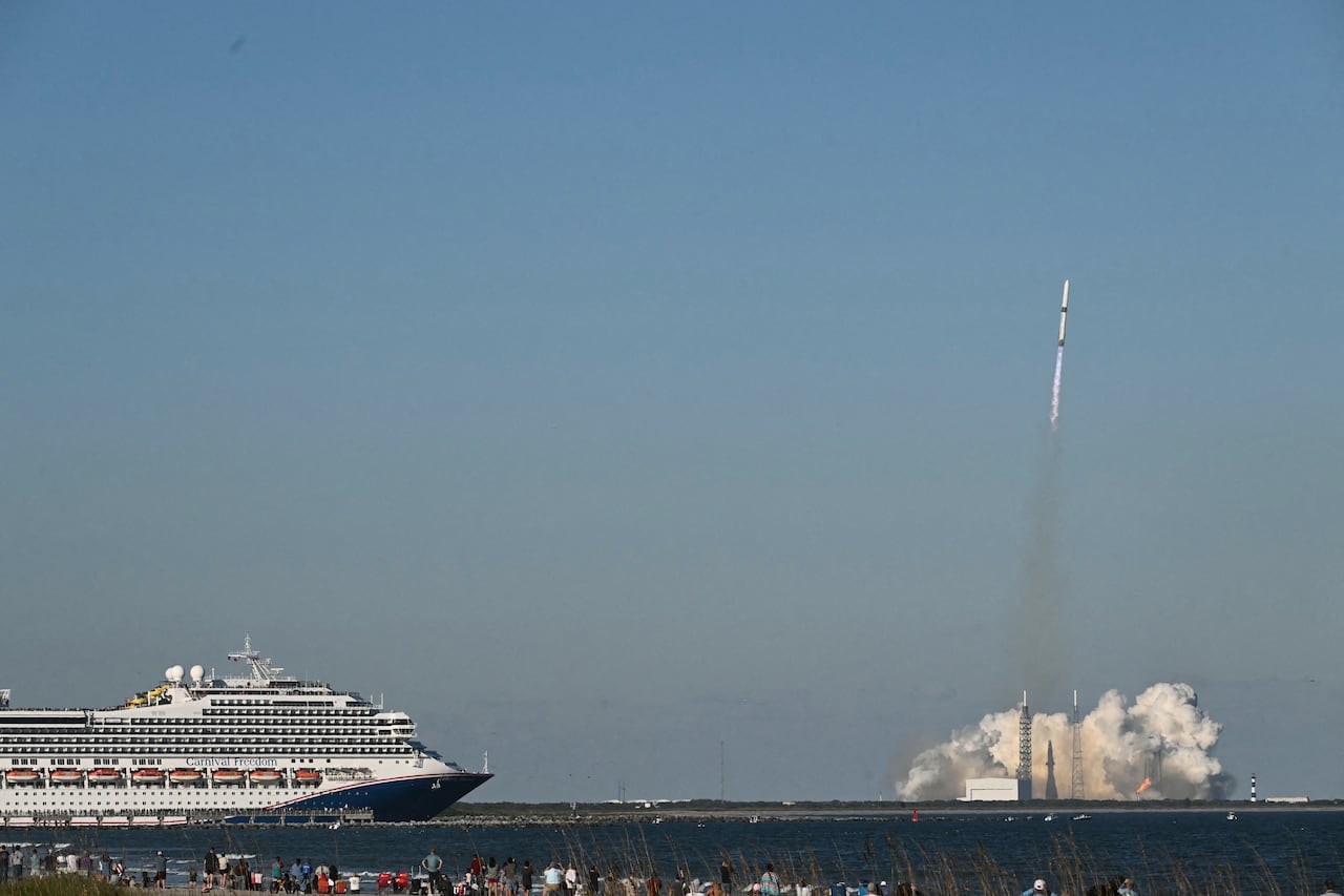 A cruise ship in the foreground, with a rocket heading for the sky seen in the background