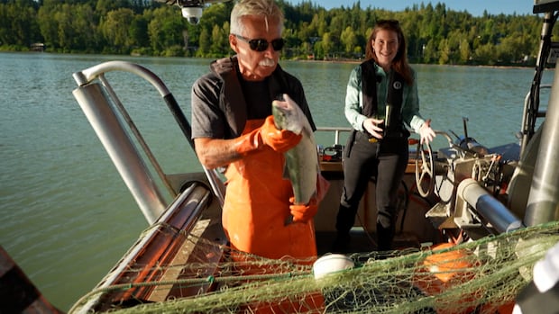A man on a boat holds a fish over a net, a woman talks in the background. 