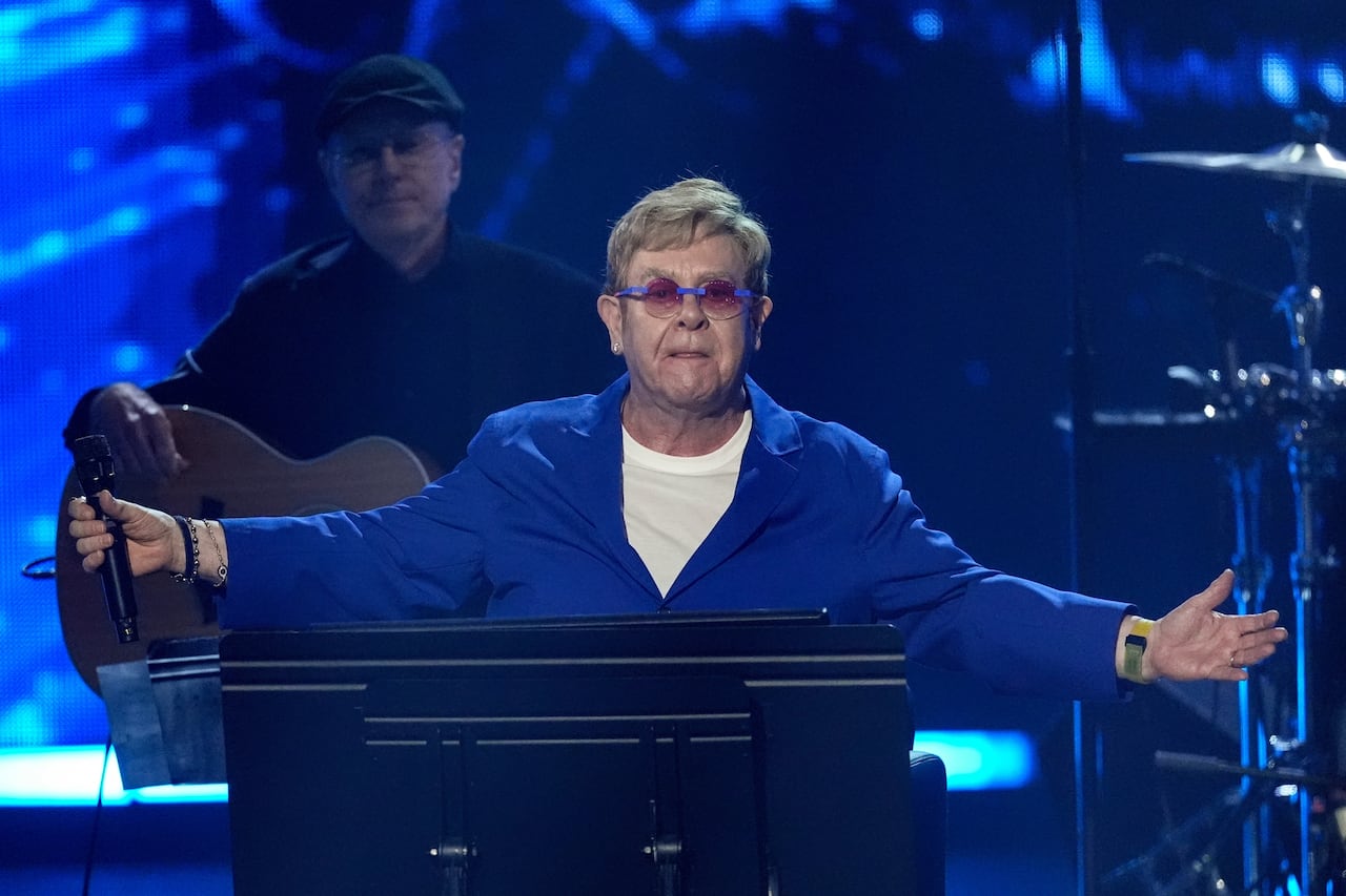 Elton John licks his lips with his arms spread wide while standing on stage