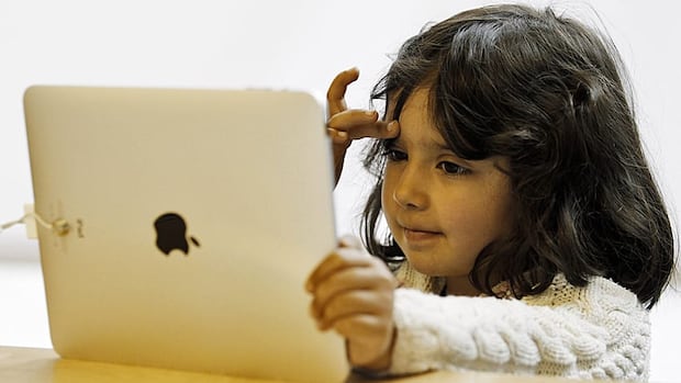 A new study by a team of researchers from two Toronto hospitals that was published in the Journal of the American Medical Association found that children who spent more time on screens before age eight scored lower on standardized tests. A young girl in a white sweater with short dark hair looks at the screen of an iPad.