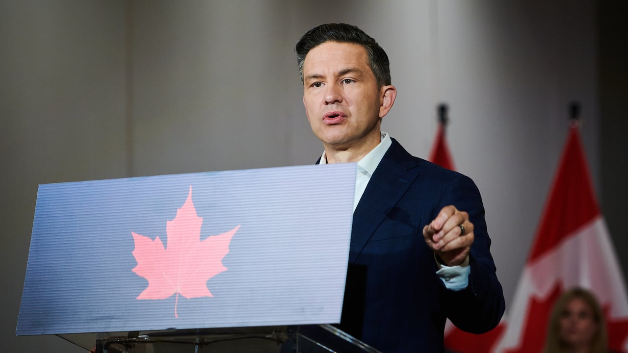 Pierre Poilievre stands at podium with Canadian flags and a woman in the background