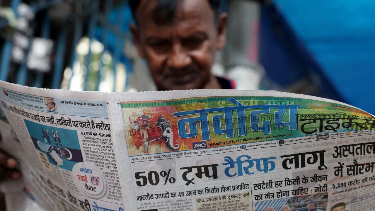 A man reads a Hindi newspaper with a colorful illustration at the top. The headline mentions a 50% tariff. The setting appears casual and focused.