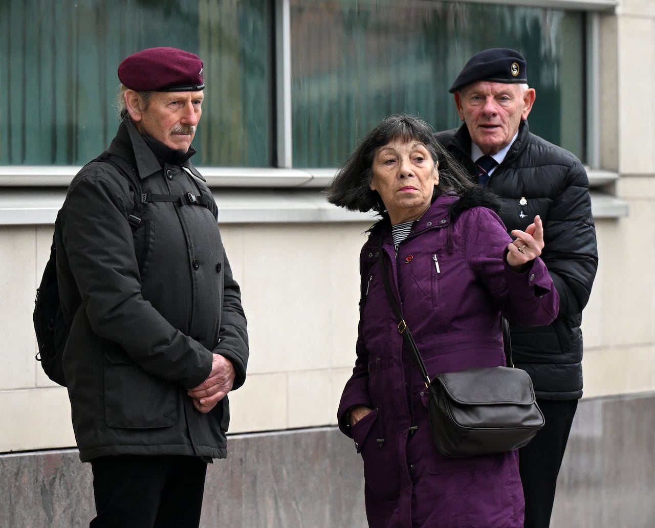 Two older men in military berets and an older woman are shown standing on a sidewalk.