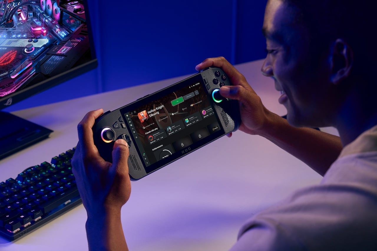 Promotional image of a man holding a portable gaming system in front of a computer desk.
