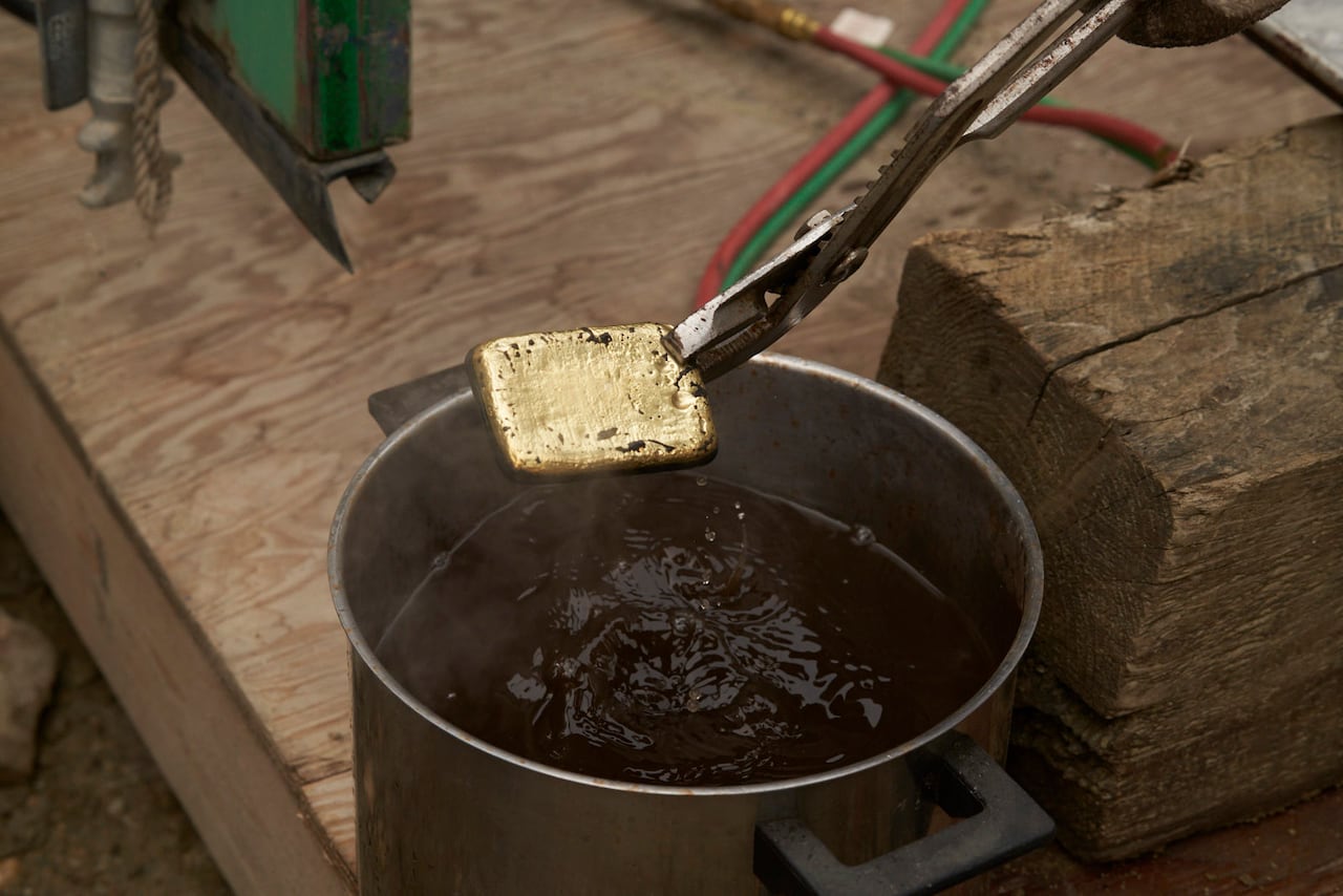 A pair of metal tongs holds a crude-looking god bar over a container of water.