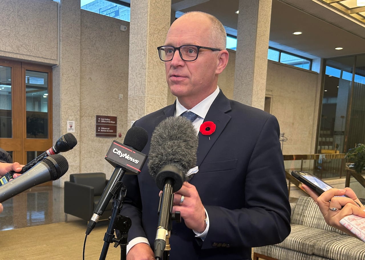 A bald man with glasses and a blue blazer speaks into several microphones