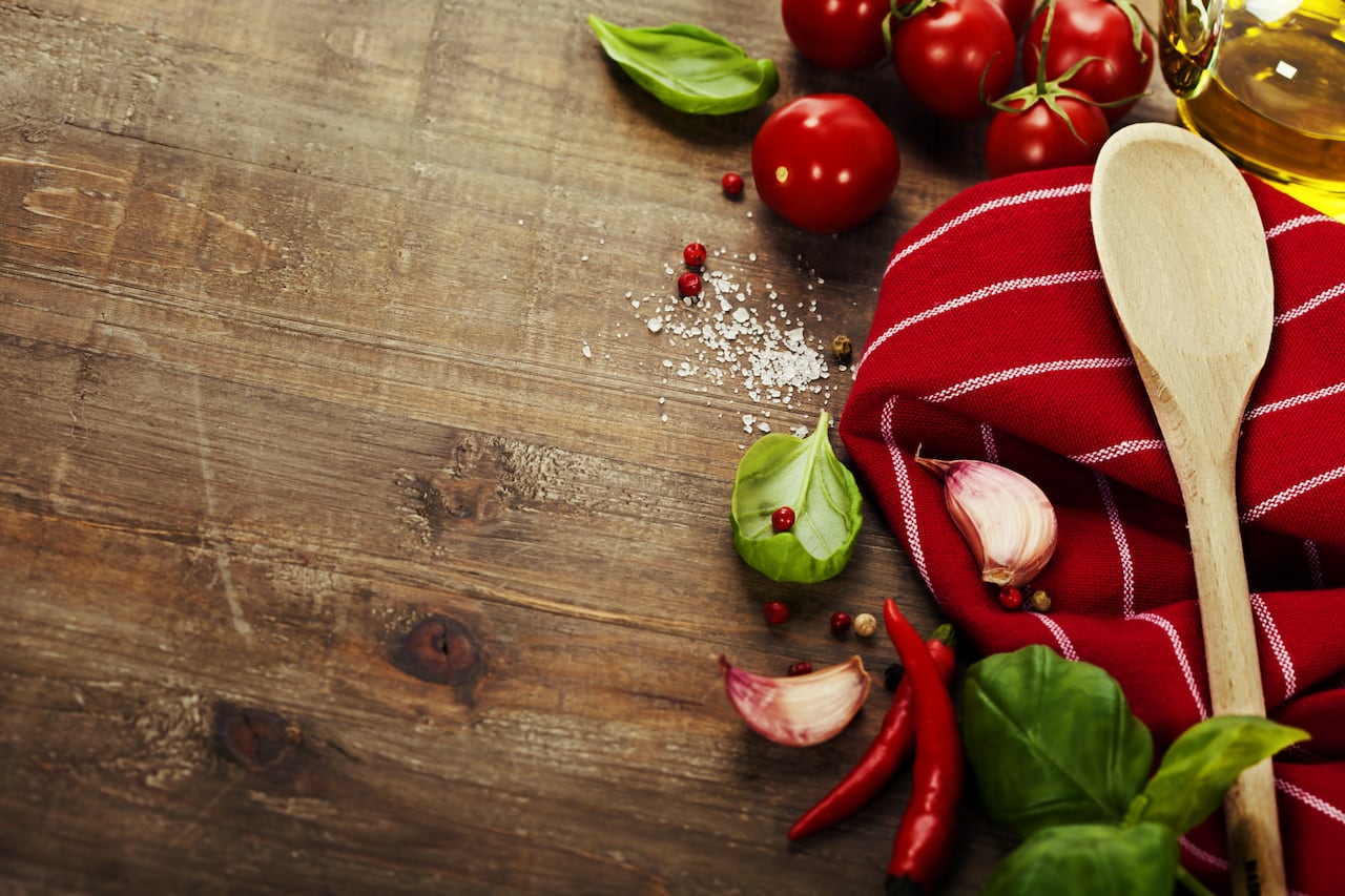 A wooden spoon and red dish towel are seen on a wooden table, with garlic, basil, tomatoes, olive oil, peppers and salt.