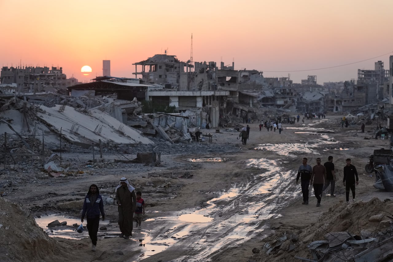 People walk along the muddy road as destroyed buildings are seen near them.