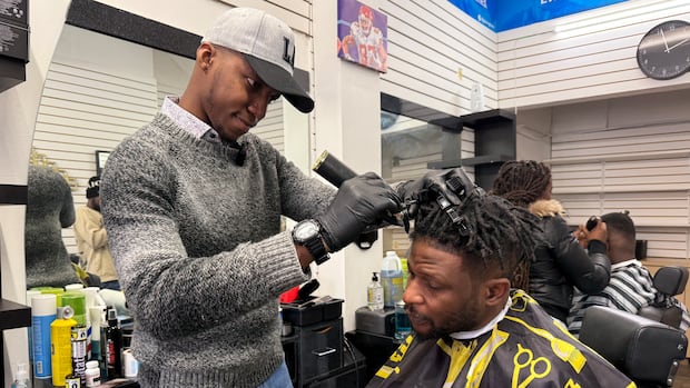 Abdul Ndikumana, left, has been gaining experience volunteering and now working at the Giving Trim barbershop in Centretown. A man in grey sweater holds an electric razor to the head of a seated man wearing a black and yellow smock.